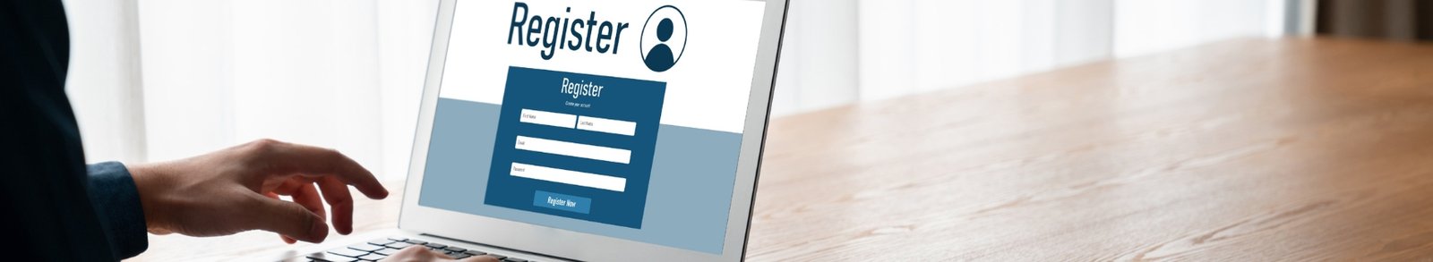 An individual is shown from a side profile, sitting at a wooden desk and actively using a silver laptop. Their hands are positioned over the keyboard and trackpad, suggesting they are in the middle of a task. The laptop screen displays a clean, professional "Register" webpage featuring a prominent user icon and a blue sign-up form with fields for first name, last name, email, and password. The background is softly blurred with bright, natural light filtering through white curtains, creating a focused and modern atmosphere for an online registration process.