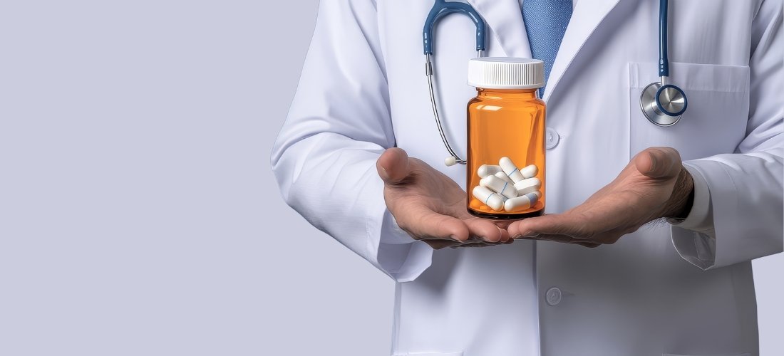 A wide banner image features a doctor in a white lab coat with a blue stethoscope around their neck, holding an amber-colored prescription bottle in the palm of their hand. The transparent bottle is filled with several white capsules. The doctor's face is not visible, focusing attention on the medication. The background is a clean, solid light grey, with ample empty space on the left side, making it ideal for healthcare-related messaging or clinical information.