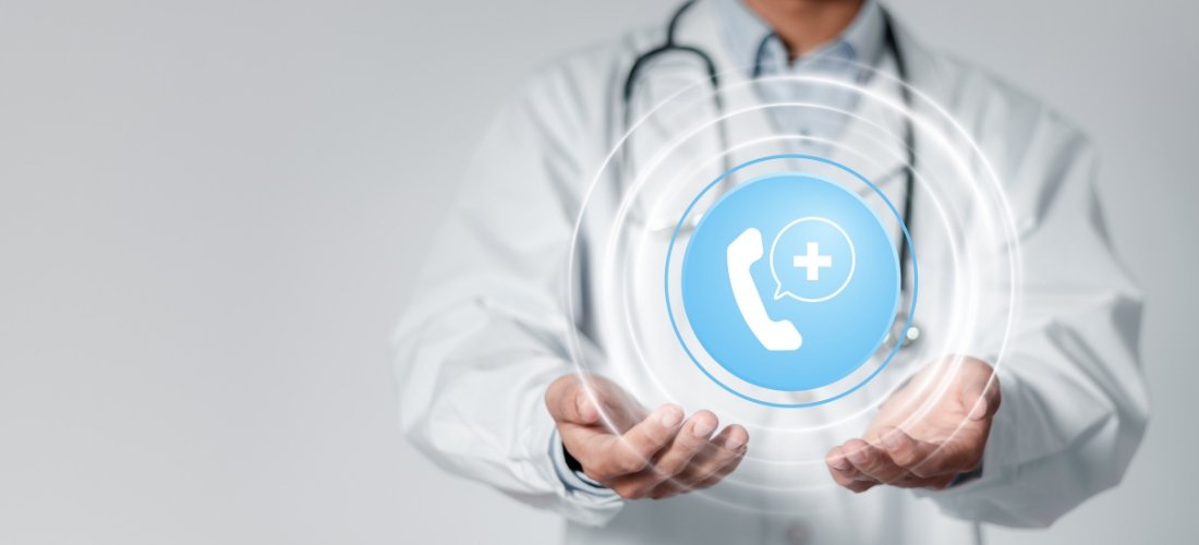 This image shows a doctor wearing a white coat and a stethoscope around their neck, holding their hands open in front of them. Above their hands is a glowing blue circular icon with a white telephone handset and a medical cross inside a speech bubble, symbolizing medical help or telehealth services. Surrounding the icon are concentric white rings, suggesting wireless communication or connectivity, emphasizing the concept of accessible healthcare through phone or digital consultation.