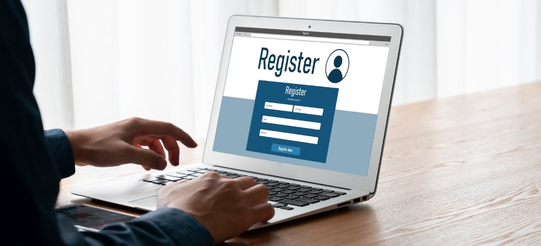 An individual is shown from a side profile, sitting at a wooden desk and actively using a silver laptop. Their hands are positioned over the keyboard and trackpad, suggesting they are in the middle of a task. The laptop screen displays a clean, professional "Register" webpage featuring a prominent user icon and a blue sign-up form with fields for first name, last name, email, and password. The background is softly blurred with bright, natural light filtering through white curtains, creating a focused and modern atmosphere for an online registration process.
