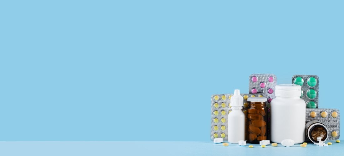 A wide banner-style image features a variety of medical supplies arranged on the right side against a solid light blue background. The collection includes several blister packs with colorful yellow, pink, and green pills, along with a white plastic pill bottle, a small amber glass bottle filled with brown capsules, and a white nasal spray bottle. A few loose white and yellow tablets are scattered at the base of the containers. The left side of the image is left entirely as open blue space, providing a clean area for text or branding. The overall composition is bright and organized, conveying a theme of healthcare, pharmacy, or medication management.